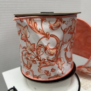 Op de afbeelding: Een rol wit lint met een herhalend oranje en perzikkleurig bloemmotief in krulvorm. Het lint heeft afgewerkte randen met een dun oranje koordje aan de boven- en onderkant en is gewikkeld rond een ronde kartonnen kern.