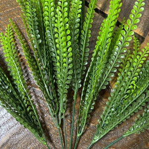 May include: A collection of artificial green grass stems. The stems are a bright green, with small, rounded leaves along the sides. The stems are grouped together, giving a full and textured look. The background is a wooden surface.