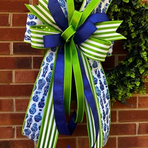 May include: A decorative bow with a green, white, and blue color scheme. The bow features a variety of ribbons, including striped and patterned designs. The background is a brick wall and a green wreath.