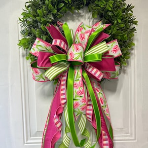 May include: A decorative wreath with a dense green base and a colourful bow. The bow is made of pink, white, and green ribbons, some patterned with tulips and others with stripes. Suitable for door or wall decoration.