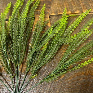 Puede incluir: Un ramo de vegetación artificial con tallos largos y delgados y hojas pequeñas y redondeadas. Las hojas cambian de un verde claro brillante en las puntas a un verde más oscuro en la base. Los tallos son de color verde oscuro.