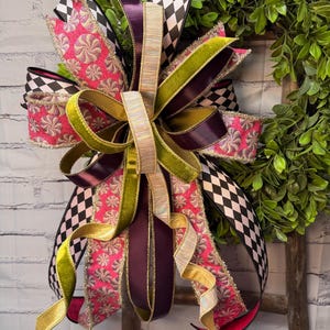 May include: A decorative bow made of multiple ribbons featuring a black and white diamond pattern, a bright pink fabric with a metallic swirl design, solid dark purple satin, olive green velvet, and a shimmering iridescent gold ribbon.