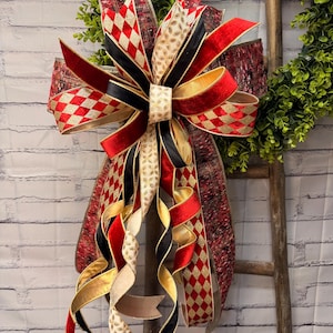 May include: A decorative bow featuring multiple ribbons in red, black, gold, and cream. The ribbons display patterns including red and gold diamonds, solid red velvet, solid black, and a cream-colored paisley print with long, curling tails.
