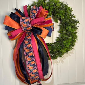 May include: A green boxwood wreath adorned with a large, multi-layered decorative bow. The bow features ribbons in navy blue, bright orange, and hot pink, with a central ribbon displaying a pattern of orange pumpkins on a dark blue background.
