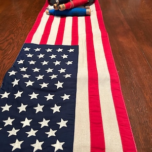 Könnte beinhalten: Ein Tischläufer im Design der amerikanischen Flagge. Der Läufer hat ein blaues Feld mit weißen Sternen und rot-weißen Streifen. Mehrere Garnrollen in Rot, Weiß und Blau liegen obenauf.