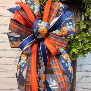 May include: A decorative bow featuring multiple ribbon styles, including a navy blue base with a pumpkin and gourd print, a solid orange ribbon, and a multi-coloured plaid ribbon. The bow has long, flowing tails and is arranged in a full, layered design.
