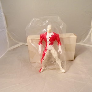 May include: A plastic action figure featuring a white and red color scheme, standing in a dynamic pose. The figure is encased in a clear plastic bag and is positioned in front of a plain, rectangular white cardboard box.