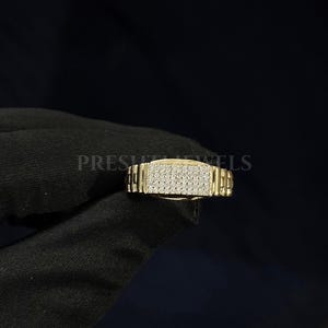 May include: A gold-colored ring with a rectangular face covered in small, sparkling stones. The band has a textured, watch-band style design. The word "PRESH" is partially visible on the left side of the ring.
