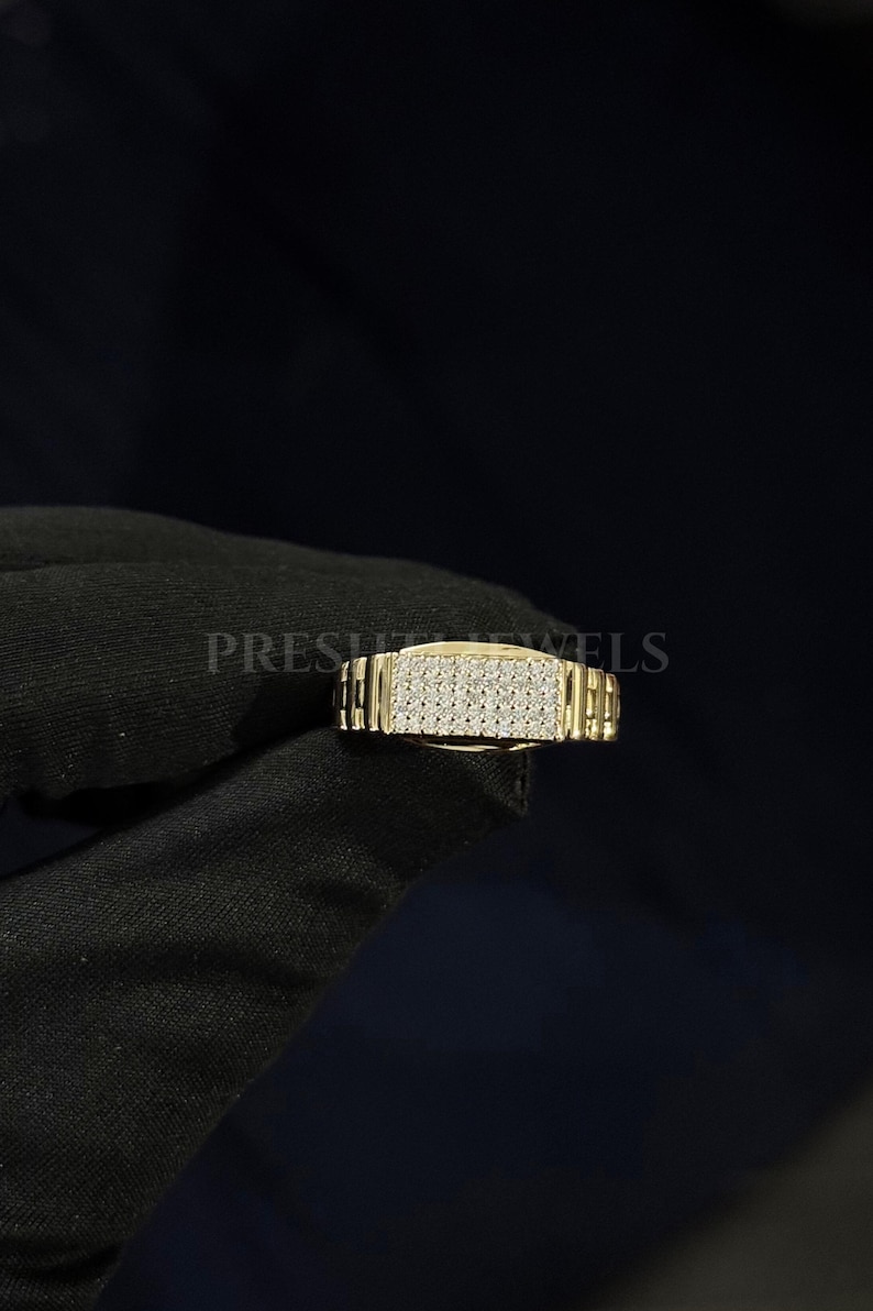 May include: A gold-colored ring with a rectangular face covered in small, sparkling stones. The band has a textured, watch-band style design. The word "PRESH" is partially visible on the left side of the ring.