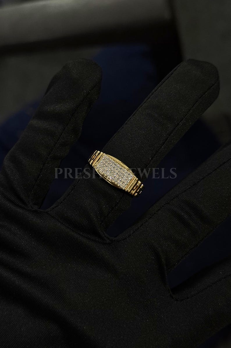 May include: A gold-colored ring with a rectangular face covered in small, sparkling stones. The band has a textured design. The word "PRES" and "WELS" are partially visible on the image.