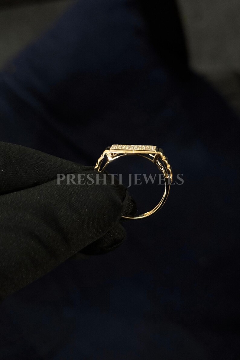 May include: A gold ring with a rectangular top encrusted with small, clear stones. The band has a delicate, rope-like design. The ring is held against a dark background. The text "PRESHTI JEWELS" is visible.
