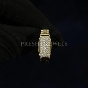 May include: A gold-colored ring with a rectangular face covered in small, sparkling stones. The band has a textured, watch-band style design. The word "PRESH JEWELS" is visible in the background.