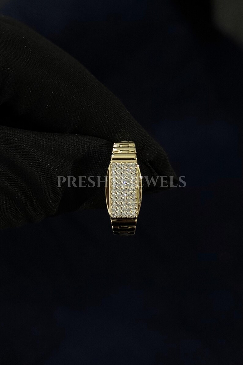 May include: A gold-colored ring with a rectangular face covered in small, sparkling stones. The band has a textured, watch-band style design. The word "PRESH JEWELS" is visible in the background.