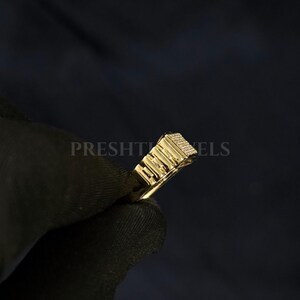 May include: A gold-colored ring with a rectangular top, featuring a textured design and small, sparkling details. The ring is held against a dark background, with the word "PRESHT" partially visible.