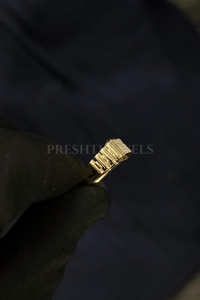May include: A gold-colored ring with a rectangular top, featuring a textured design and small, sparkling details. The ring is held against a dark background, with the word "PRESHT" partially visible.