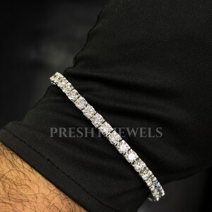 May include: A tennis bracelet with numerous round-cut diamonds. The bracelet is displayed on a black sleeve. The text "PRESH JEWELS" is visible on the sleeve. The diamonds sparkle, reflecting light.