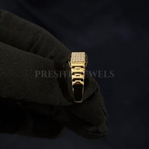 May include: A gold-colored ring with a rectangular top encrusted with small, sparkling stones. The ring has a detailed, geometric design on the sides and is held against a dark background. The text "PRESHT JEWELS" is visible.