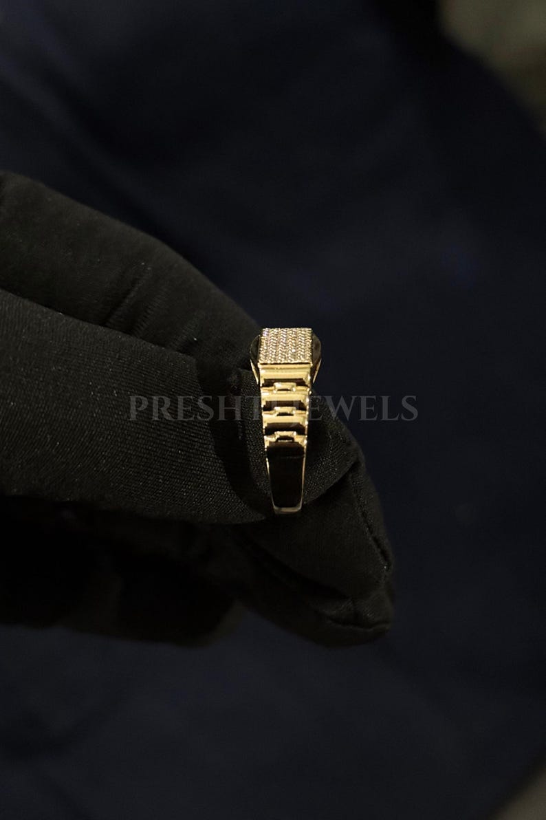 May include: A gold-colored ring with a rectangular top encrusted with small, sparkling stones. The ring has a detailed, geometric design on the sides and is held against a dark background. The text "PRESHT JEWELS" is visible.