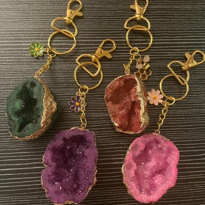 May include: Keyrings with colourful geode pendants and gold-coloured trim. Each keyring has a gold-coloured clasp and decorative charms, including flowers and a bee. The geodes are in shades of green, purple, red, and pink.