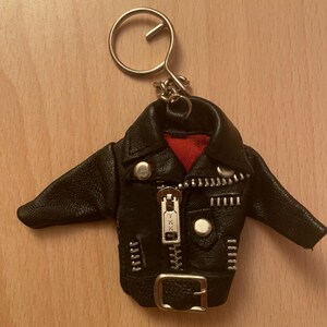 May include: A small black leather jacket keyring with silver-coloured metal hardware, including a zip, studs, and a buckle. The jacket has a red lining and is attached to a metal keyring.