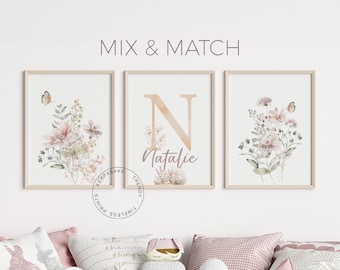 May include: Three framed prints with a watercolor floral design. The center print features the letter "N" in a gold color with the name "Natalie" written below it. The prints are hung on a white wall above a couch with pink and white pillows.