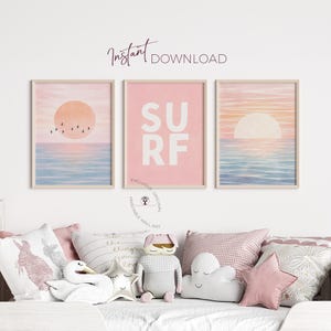 Op de afbeelding: Drie ingelijste wandkunstprints met een strandthema. De prints tonen aquarel zonsondergangen, oceaangolven en het woord "SURF" in grote witte letters op een roze achtergrond. De prints zijn in neutraal getinte houten frames.