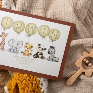 May include: A framed print featuring the name "IRWIN" in large letters on balloons, with watercolor illustrations of a giraffe, elephant, lion cub, panda, and zebra. The print includes baby's birth details. A wooden rattle is in the image.