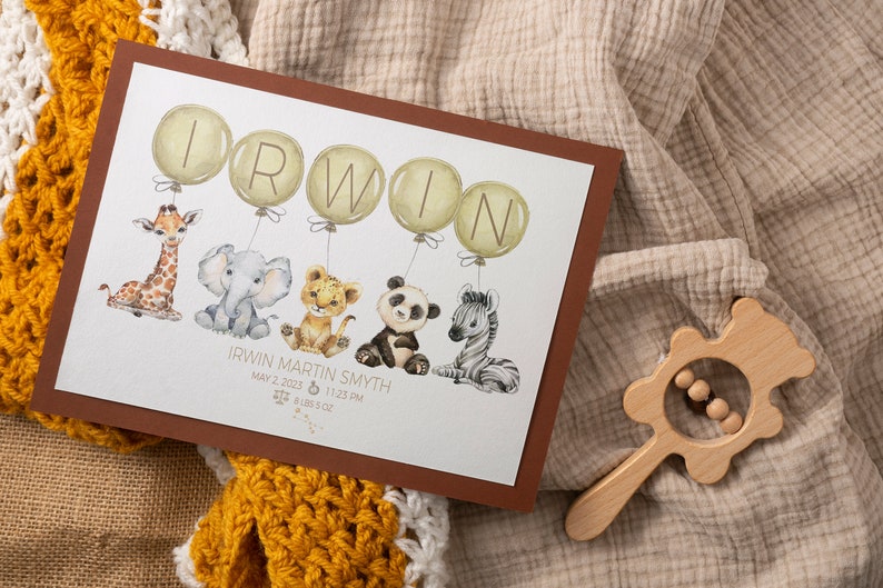 May include: A framed print featuring the name "IRWIN" in large letters on balloons, with watercolor illustrations of a giraffe, elephant, lion cub, panda, and zebra. The print includes baby's birth details. A wooden rattle is in the image.