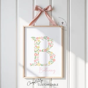 May include: A framed floral print featuring the letter "B" composed of pink, yellow, and green flowers. The name "Bethany" is written in a soft pink script below the letter. A pink ribbon is tied at the top, and the print is hanging on a white wall.