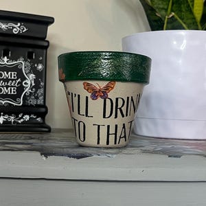 May include: A small, hand-painted flower pot with the text "I'll Drink to That" and a butterfly illustration. The rim is painted green. A black decorative box with the words "Home Sweet Home" is to the left, and a white potted plant is to the right.