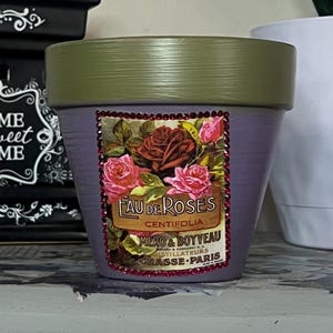 May include: A lavender-colored flower pot with a green rim. The pot features a decorative label with the words "Eau de Roses" and a floral design. The label is framed with red rhinestones. A charming addition to any home decor.