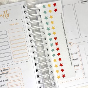 May include: Open spiral-bound planner with sections for tracking goals and results. Includes a sticker sheet with colorful star and triangle stickers. The planner has sections for water intake, calories, and exercise sessions.