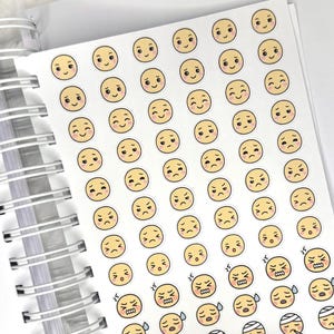 May include: A sheet of stickers featuring various emoji faces with different expressions. The emojis are yellow with black eyes and mouths, depicting emotions from happy to sad, angry, and sick. The stickers are on a white sheet, with a spiral notebook binding visible.