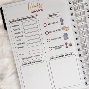May include: A white spiral-bound planner open to a "Weekly Reflection" page. The page includes sections for tracking calorie intake, water intake, and workout sessions. The text "Goals Hit" is visible, along with illustrations of a water bottle, food, protein powder, and dumbbells.