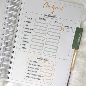 May include: Open spiral-bound planner with a white interior featuring a "Checkpoint 1" page. The page includes sections for recording measurements and updated goals. A green and gold bookmark is inserted, labeled "Checkpoint 1".