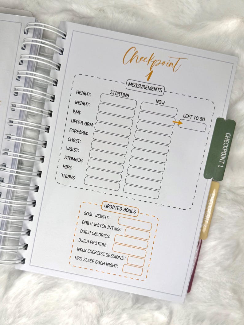 May include: Open spiral-bound planner with a white interior featuring a "Checkpoint 1" page. The page includes sections for recording measurements and updated goals. A green and gold bookmark is inserted, labeled "Checkpoint 1".