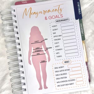 May include: A spiral-bound planner with a pink silhouette and the words "Measurements & Goals." The planner includes sections for recording body measurements and goals, with tabs labeled "Today" and "Start/Goals."