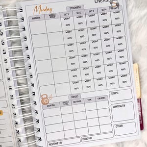 May include: Open spiral-bound fitness planner with a white interior. The planner features sections for strength training, cardio, and steps, with spaces to record exercises, weight, reps, and other metrics. The word "Monday" is written at the top.