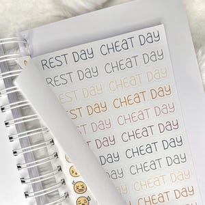 May include: A sheet of planner stickers with the phrases "REST DAY" and "CHEAT DAY" in various colors. The stickers are on a white background, with a spiral-bound notebook visible in the background. The stickers also include small emoji faces.