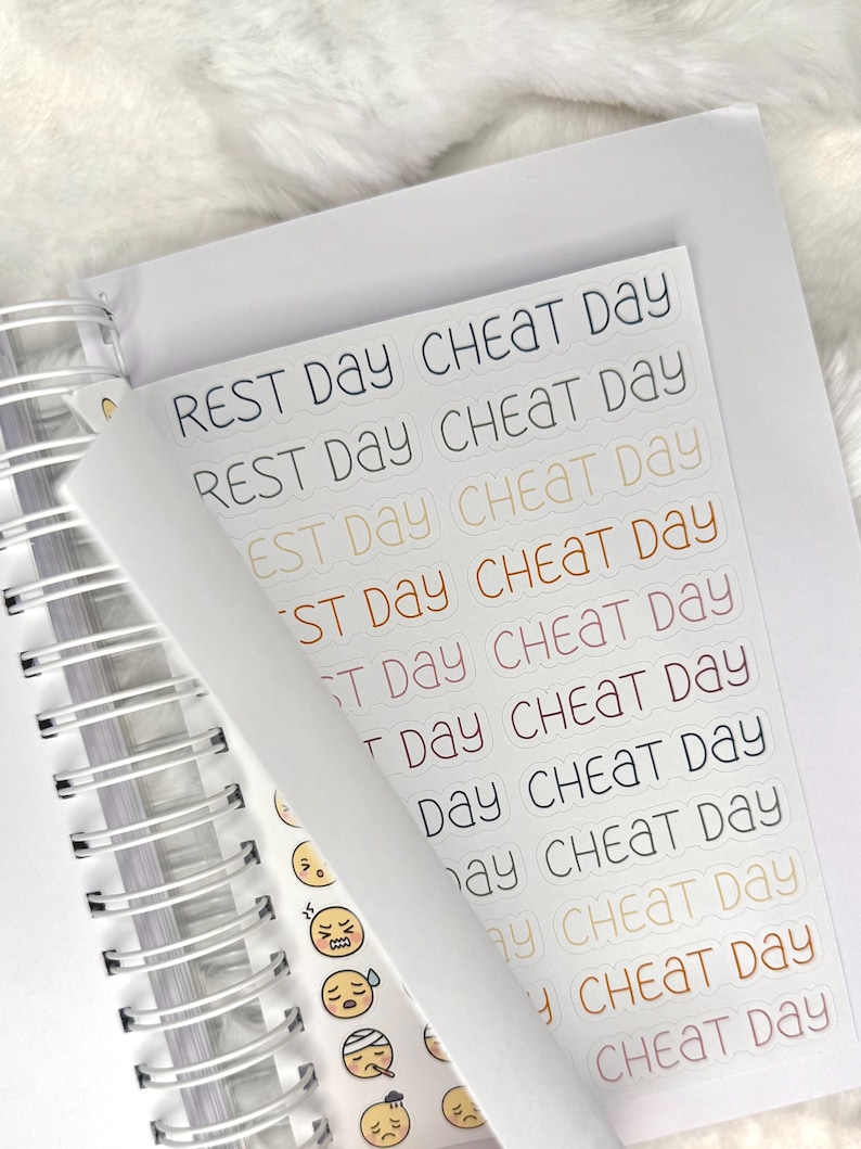 May include: A sheet of planner stickers with the phrases "REST DAY" and "CHEAT DAY" in various colors. The stickers are on a white background, with a spiral-bound notebook visible in the background. The stickers also include small emoji faces.