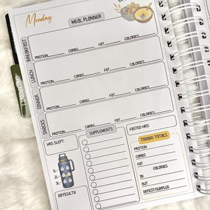 May include: A spiral-bound meal planner open to a Monday page. The page includes sections for breakfast, lunch, dinner, and snacks, with spaces for protein, carbs, fat, and calories. A water bottle graphic is on the bottom left.