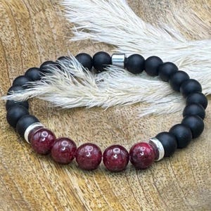 Könnte beinhalten: Ein Armband mit abwechselnd mattschwarzen und tiefroten Edelsteinperlen. Das Armband hat silberfarbene Metallakzente und ist auf einem elastischen Band aufgezogen. Das Armband wird auf einer Holzoberfläche präsentiert.