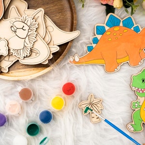 May include: Wooden dinosaur cutouts and paint pots with various colors. The orange dinosaur has blue spikes, and the green dinosaur has a yellow outline. A wooden tray holds more dinosaur shapes. A paintbrush is next to a palm tree cutout.