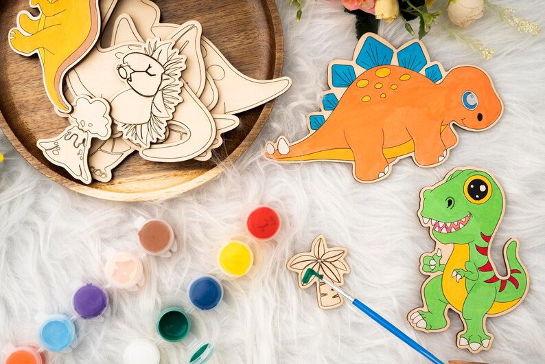 May include: Wooden dinosaur cutouts and paint pots with various colors. The orange dinosaur has blue spikes, and the green dinosaur has a yellow outline. A wooden tray holds more dinosaur shapes. A paintbrush is next to a palm tree cutout.