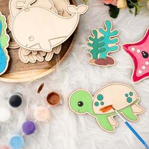 May include: A set of wooden sea creature shapes for painting, including a whale, seahorse, starfish, turtle, and seaweed. The shapes are on a white fuzzy surface with paint pots and a paintbrush.