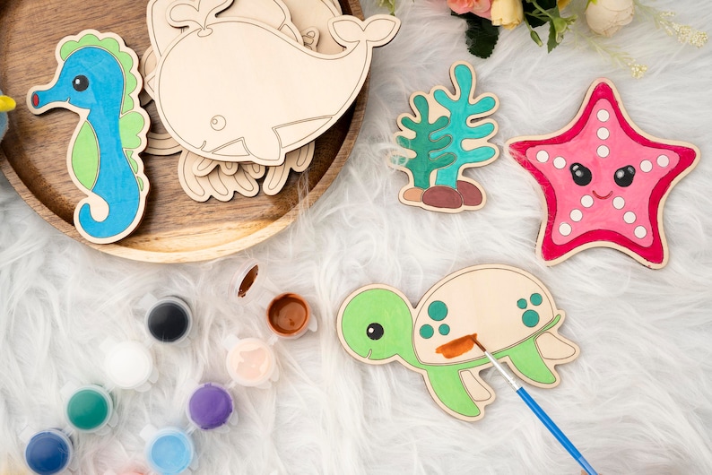 May include: A set of wooden sea creature shapes for painting, including a whale, seahorse, starfish, turtle, and seaweed. The shapes are on a white fuzzy surface with paint pots and a paintbrush.