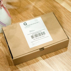 May include: A brown cardboard box with a white label and a barcode. The label includes the text "USPS GROUND ADVANTAGE". The box is on a wooden surface, next to a white vase with a red flower.