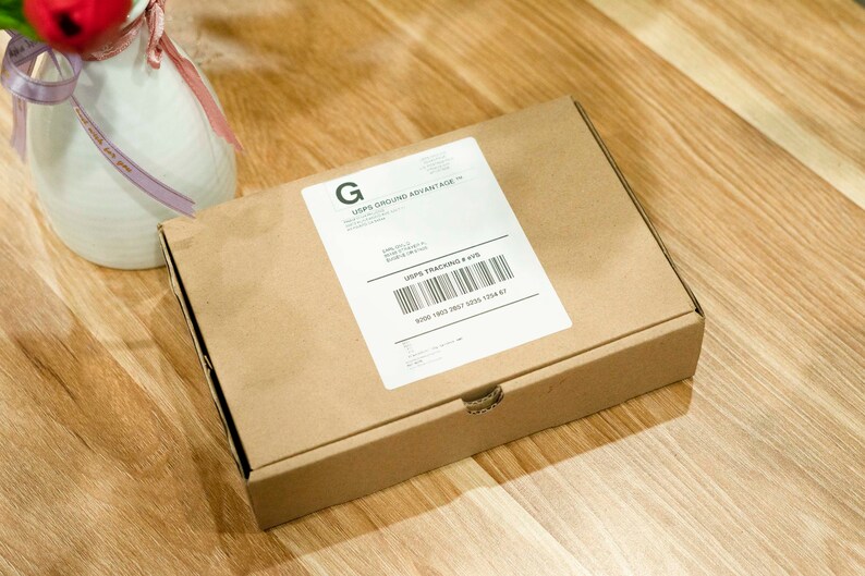 May include: A brown cardboard box with a white label and a barcode. The label includes the text "USPS GROUND ADVANTAGE". The box is on a wooden surface, next to a white vase with a red flower.