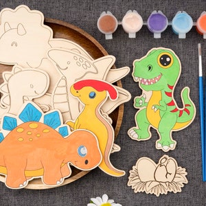 May include: A wooden tray holds various wooden dinosaur cutouts, some painted with bright colors. Paint pots and brushes are arranged nearby, suggesting a craft kit for children. The dinosaurs include a T-Rex, Stegosaurus, and others.