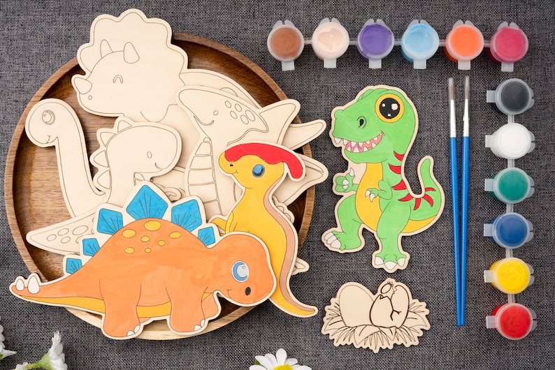 May include: A wooden tray holds various wooden dinosaur cutouts, some painted with bright colors. Paint pots and brushes are arranged nearby, suggesting a craft kit for children. The dinosaurs include a T-Rex, Stegosaurus, and others.